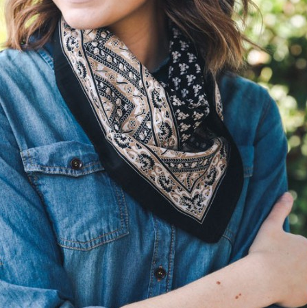 Pictured is a photo of a woman wearing a denim top and bandana scarf around her neck. The scarf is black and features tan and white floral vintage motif throughout.
