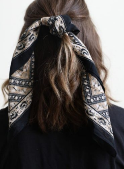 Pictured is the back of a brunette model's hair. Her hair is worn half up and tied with a black, white, and light tan scarf bandana that features floral motif throughout.