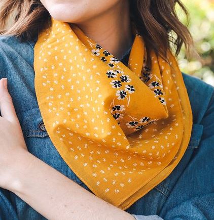 Pictured is a model wearing a denim blue top and a mustard yellow scarf around her neck. The scarf features white and blue floral motif throughout.