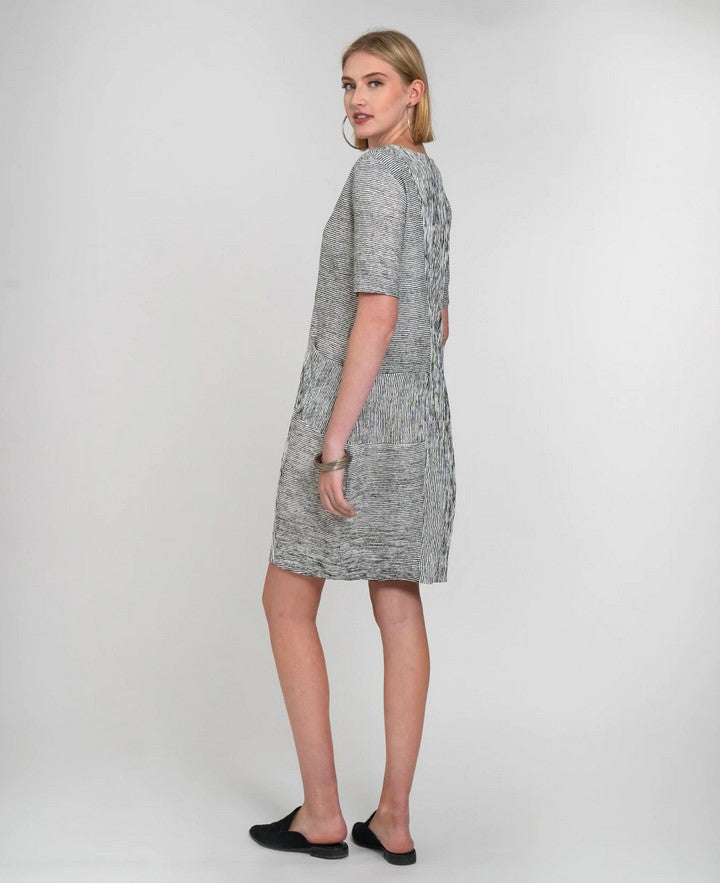 side view of blond model in a white dress with thin black stripes wearing black pointy flat mules in front of a white background