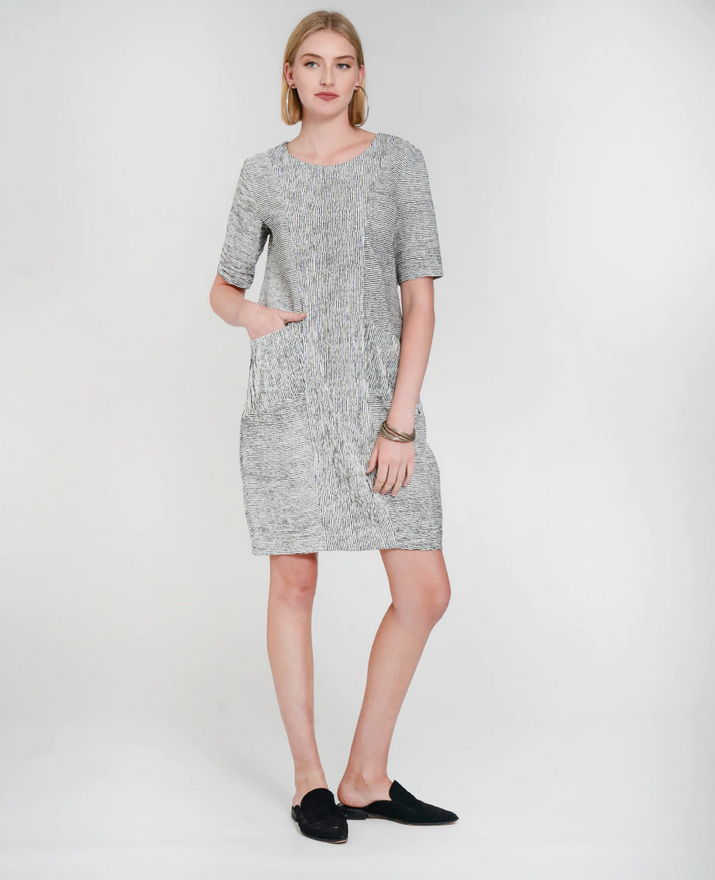 blond model in a white dress with thin black stripes wearing black pointy flat mules in front of a white background