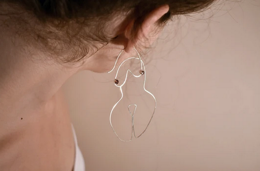 Silver wired earrings in the form a woman's body on the ears of a caucasian model.