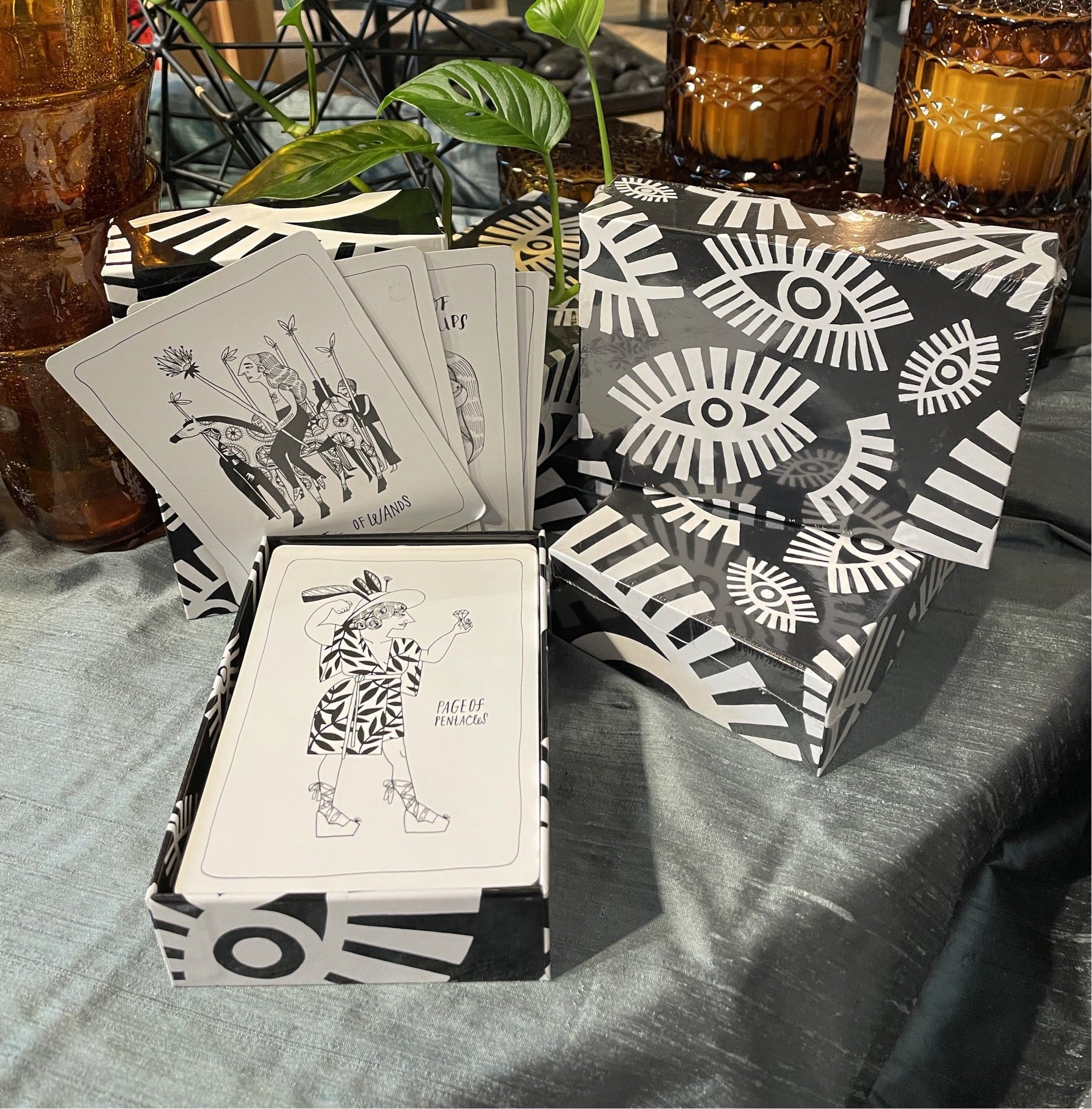 black and white evil eye pattern tarot card deck on a table with grey shiny table cloth and plants and amber glads candles in the background