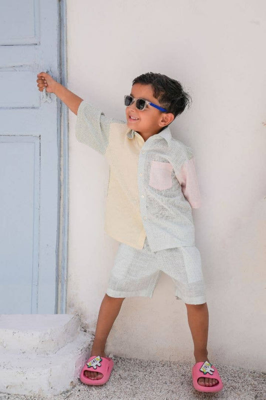 Child wearing sunglasses and a light-colored outfit standing against a white wall.
