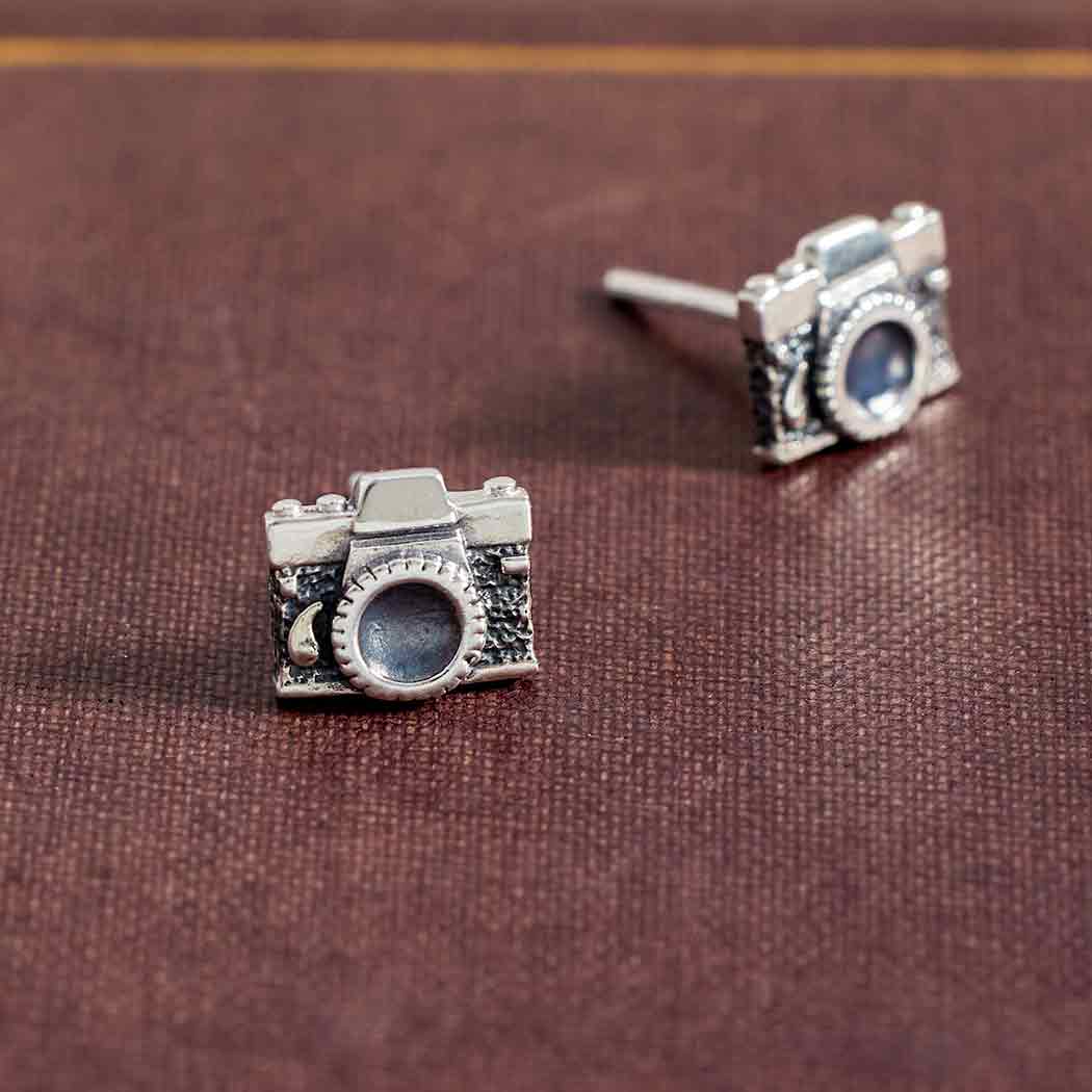 Silver camera-shaped earrings on a brown fabric background