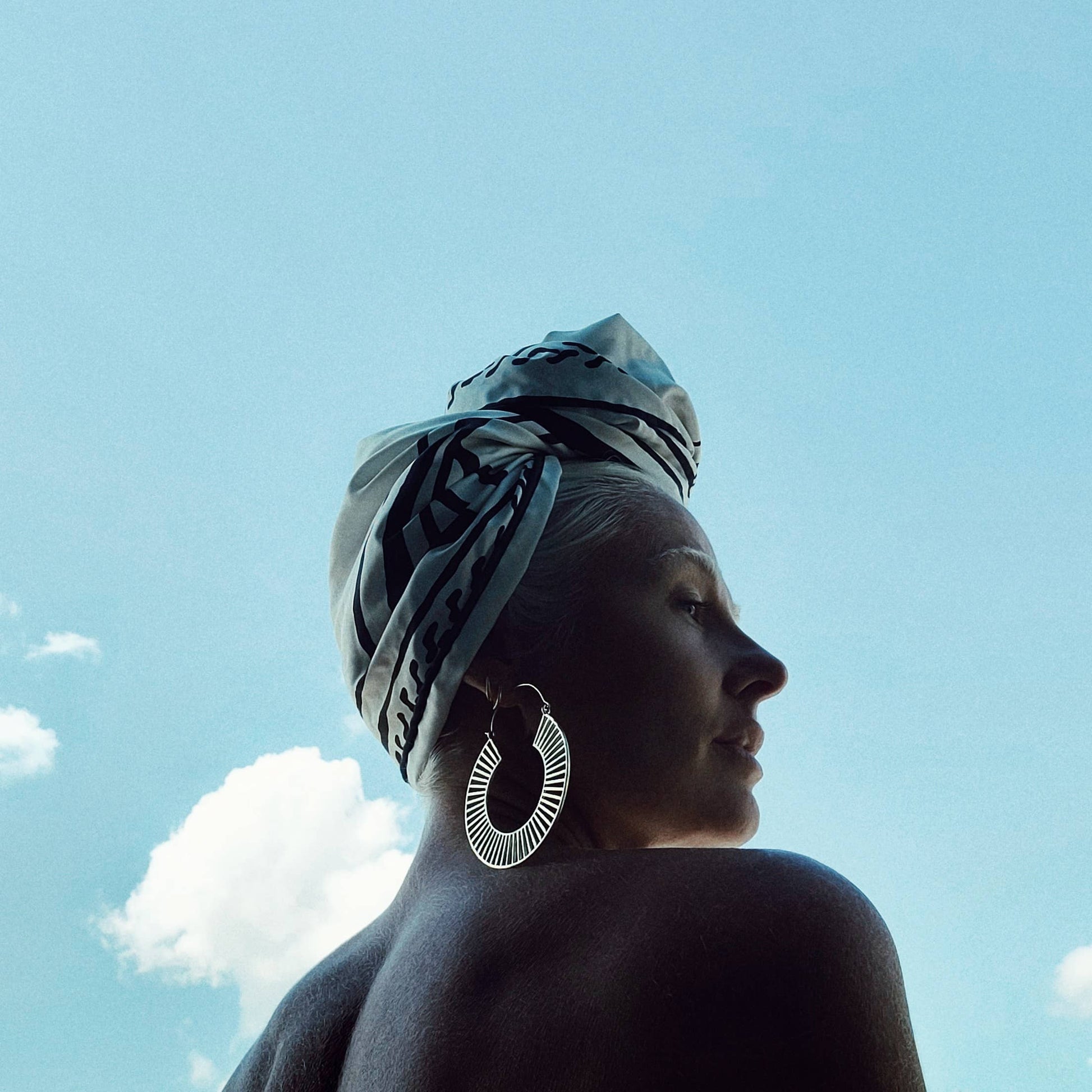 brass hoop earrings with hook closure and spindle design on model with print head wrap and blue sky with clouds