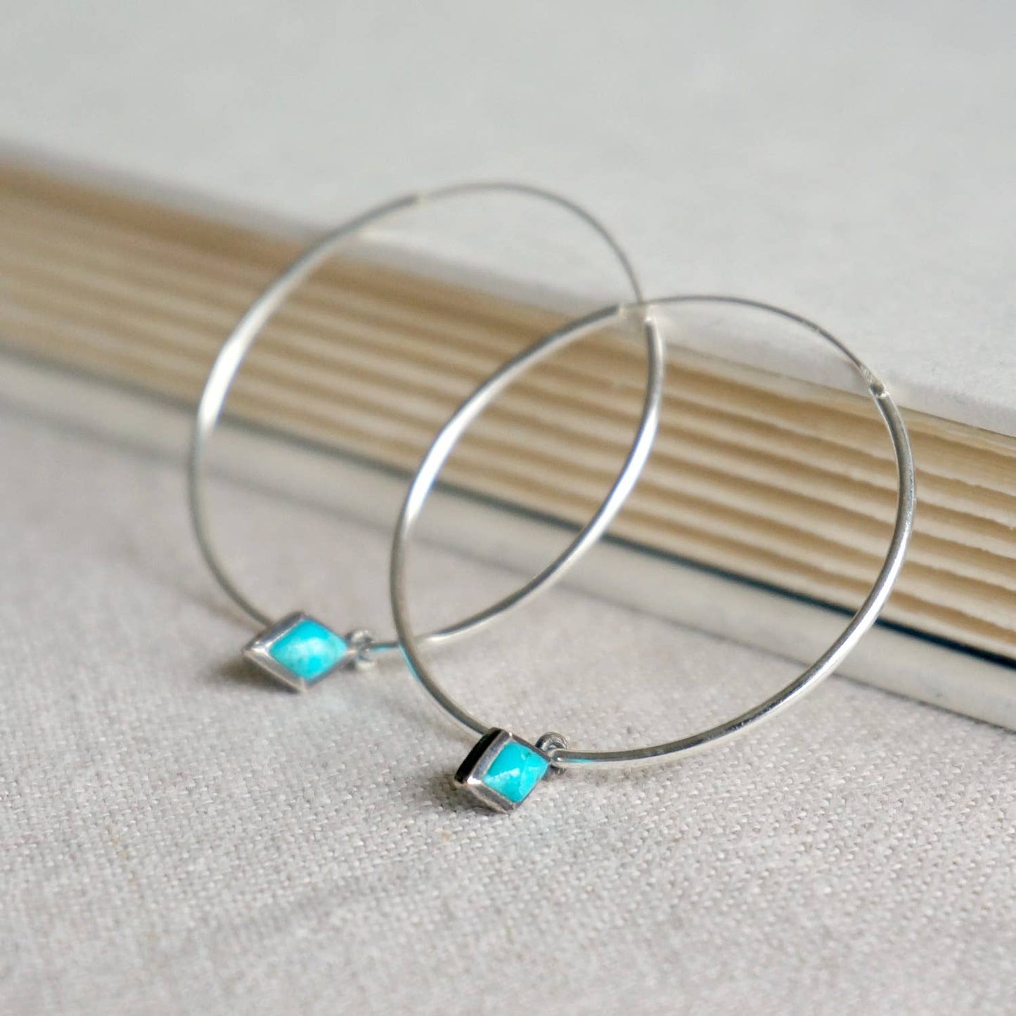 Silver hoop earrings with turquoise square stones on a textured surface