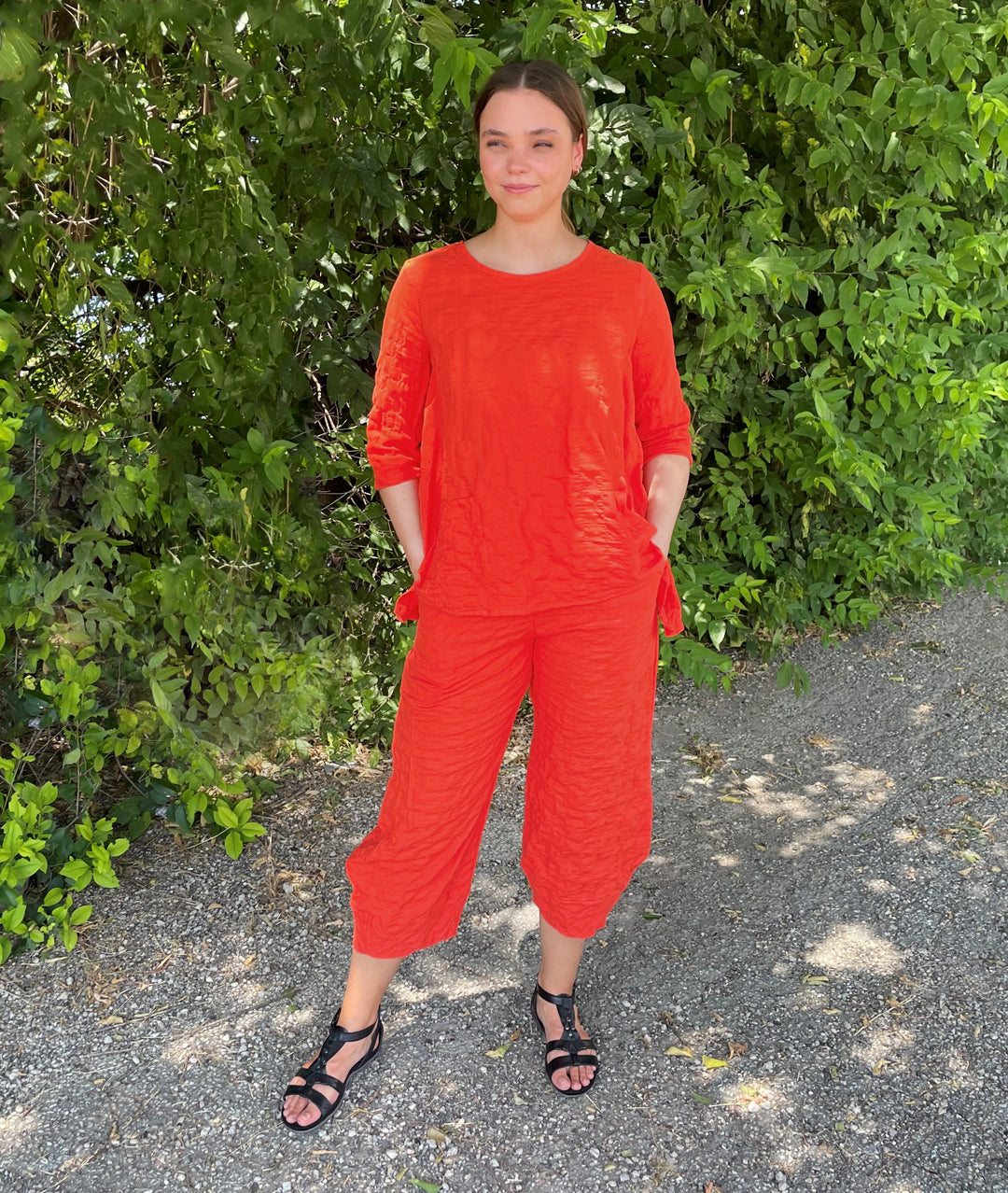 model in a chili red wide leg pant with a tapered ankle, worn with a matching pullover top with a 3/4 sleeve and dips in the hem at either hip