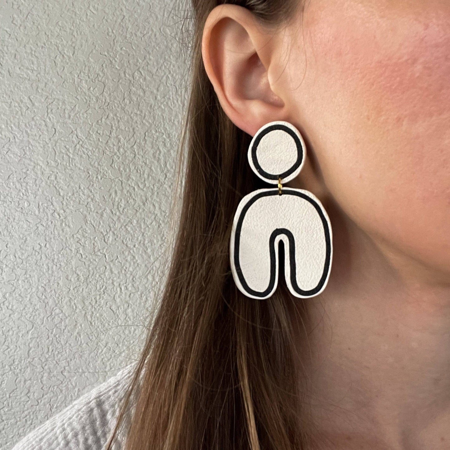 Black and white Post and dangle earrings with round port and u shaped dangle. Close up on brown haired model and white wall