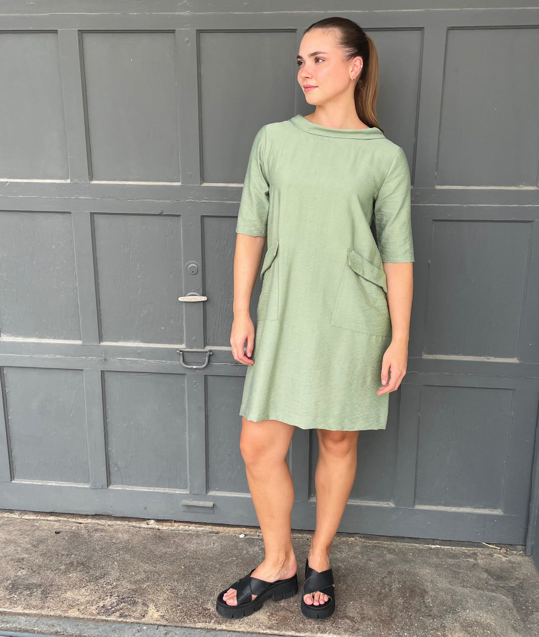 model in a moss green knee length shift dress with elbow length sleeves, a round cowl neck collar and exterior hip pockets