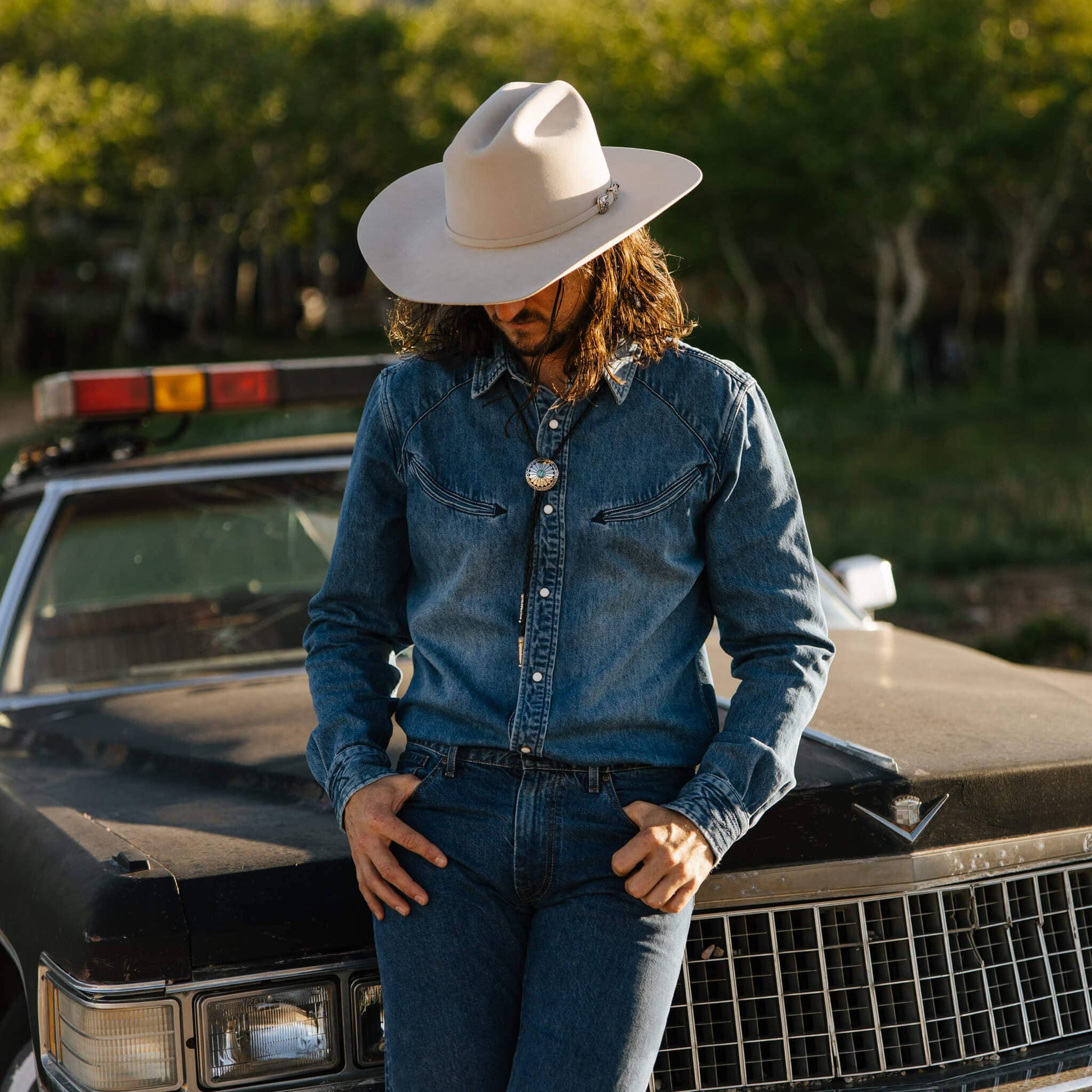 model leaning on car wearing idigo denim collared long sleeve, with arrow stitching pockets and pearl snaps. worn with jeans, beige hat and bolo tie.