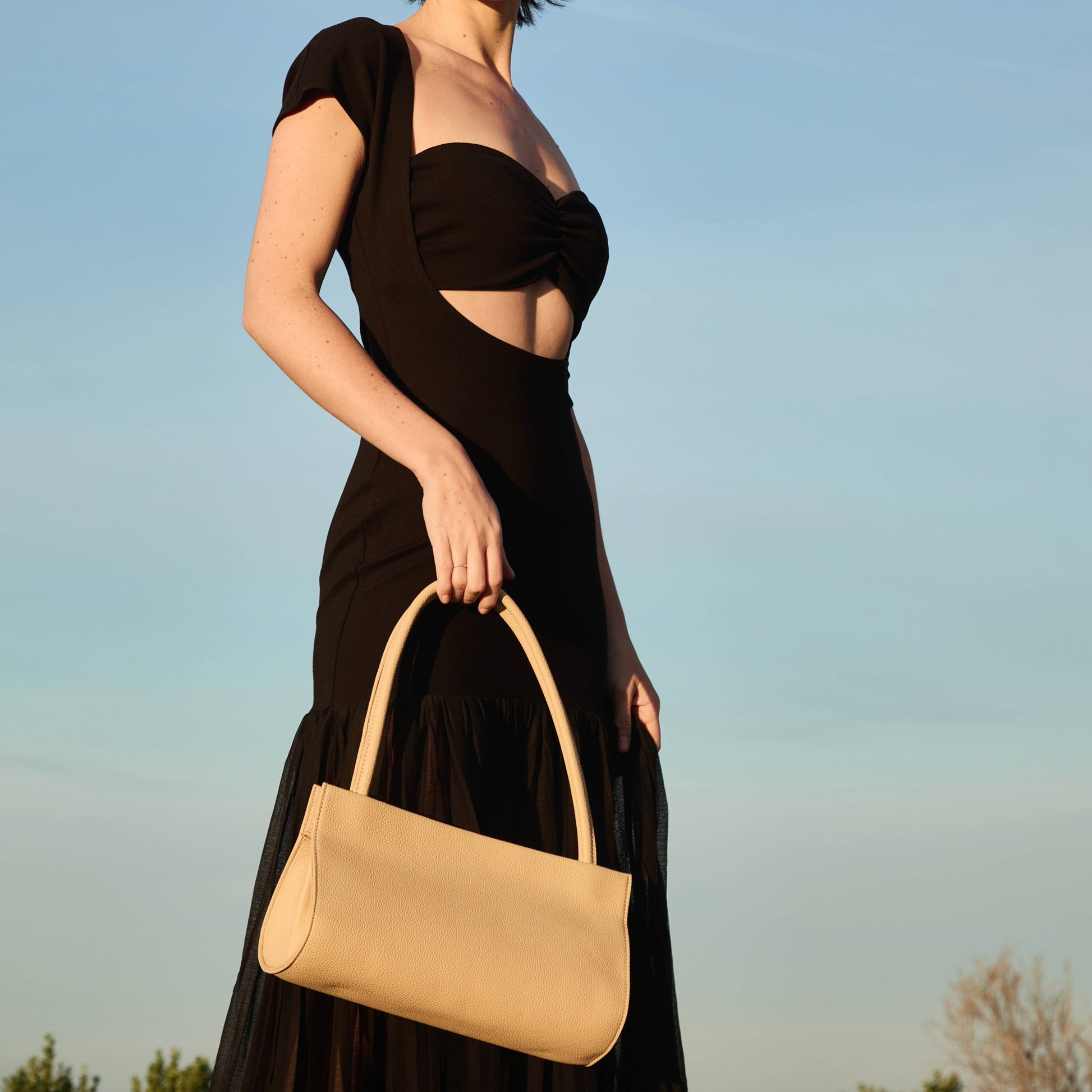 Woman in a black dress holding a beige handbag against a clear blue sky.