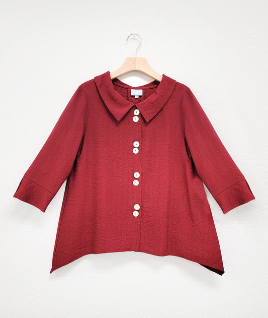 deep brick red button down bouse with four sets of double buttons, a rounded collar, 3/4 sleeves and deep dips in the side hem