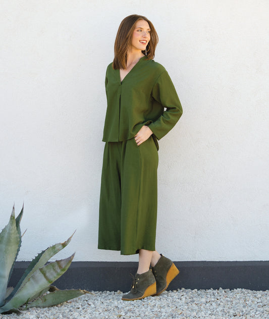 Woman in a green outfit standing against a white wall with a plant in the foreground.