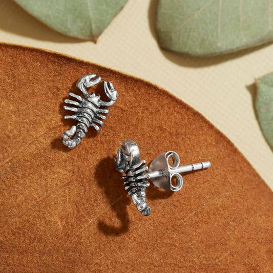 A pair of sterling silver scorpion earrings displayed on a brown textured surface with a few green leaves scattered around.