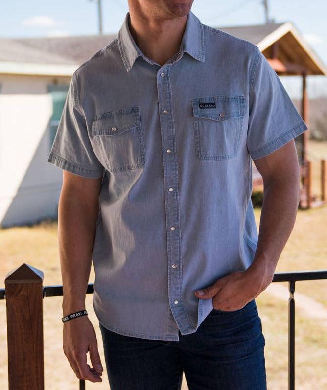 male model wearing short sleeve light blue denim pearl snap shirt in front of a fence and building 