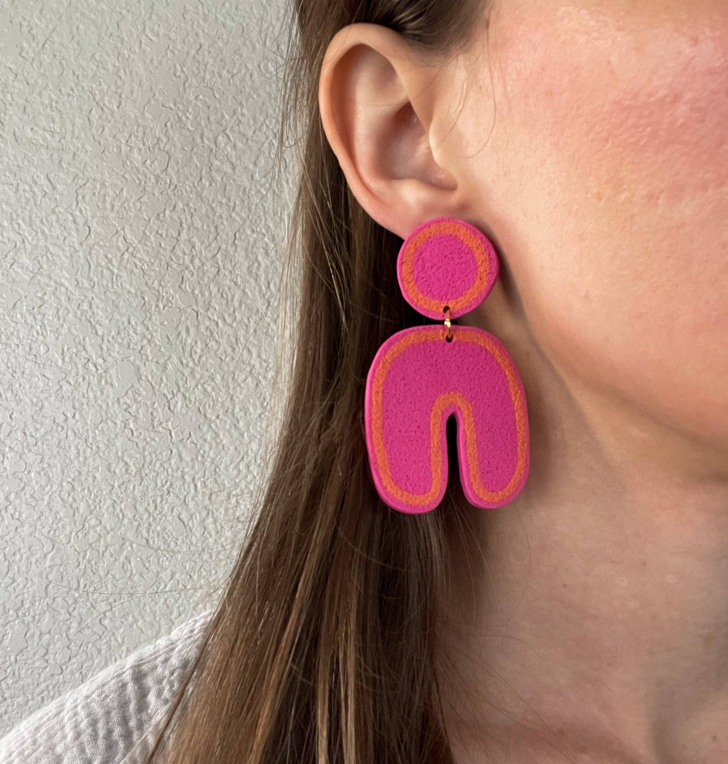 Pink and orange Post and dangle earrings with round port and u shaped dangle. White wall background adn close up of brunette model