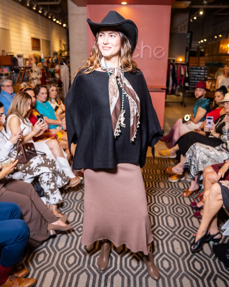 model wearing a black cowboy hat, black poncho, knit taupe skirt, brown scarf, long turquoise beaded moon necklace and boots walking down a runway