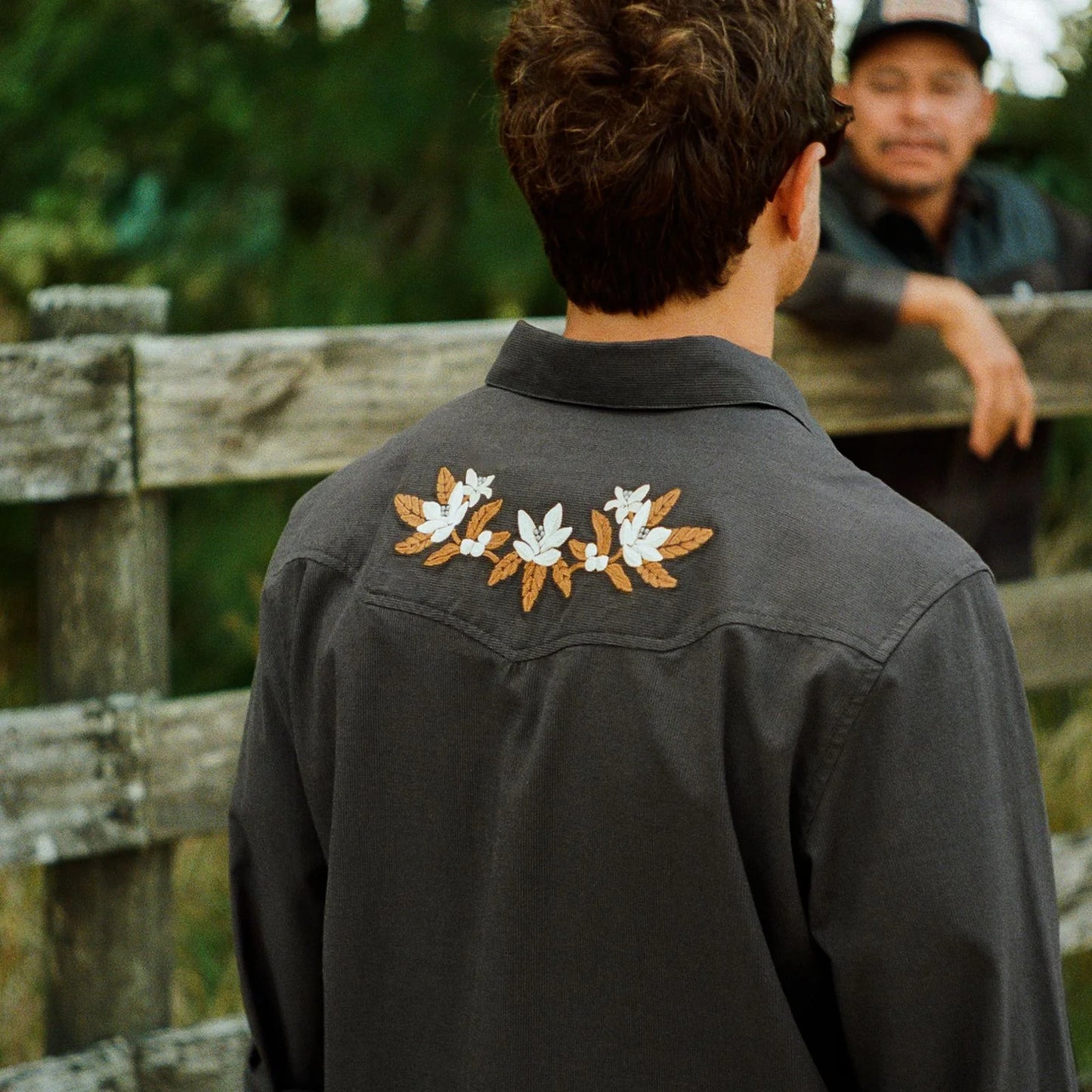 A man viewed from behind wearing a dark charcoal gray button-up shirt with embroidered white flowers and brown leaves on the back collar. He is standing outdoors in front of a rustic wooden fence, with another man visible in the blurred background.