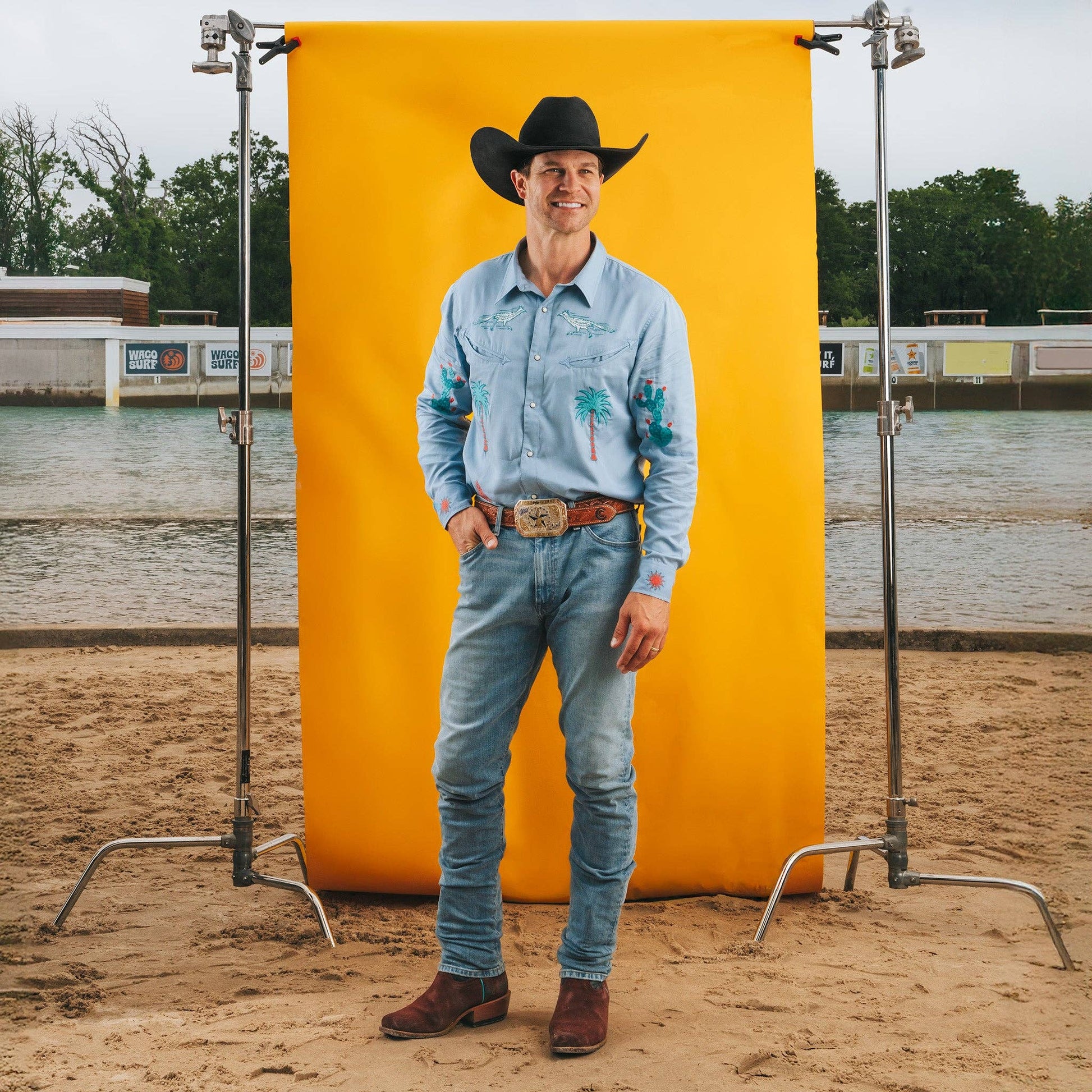 Man in cowboy attire standing in front of a yellow backdrop at a rodeo arena.