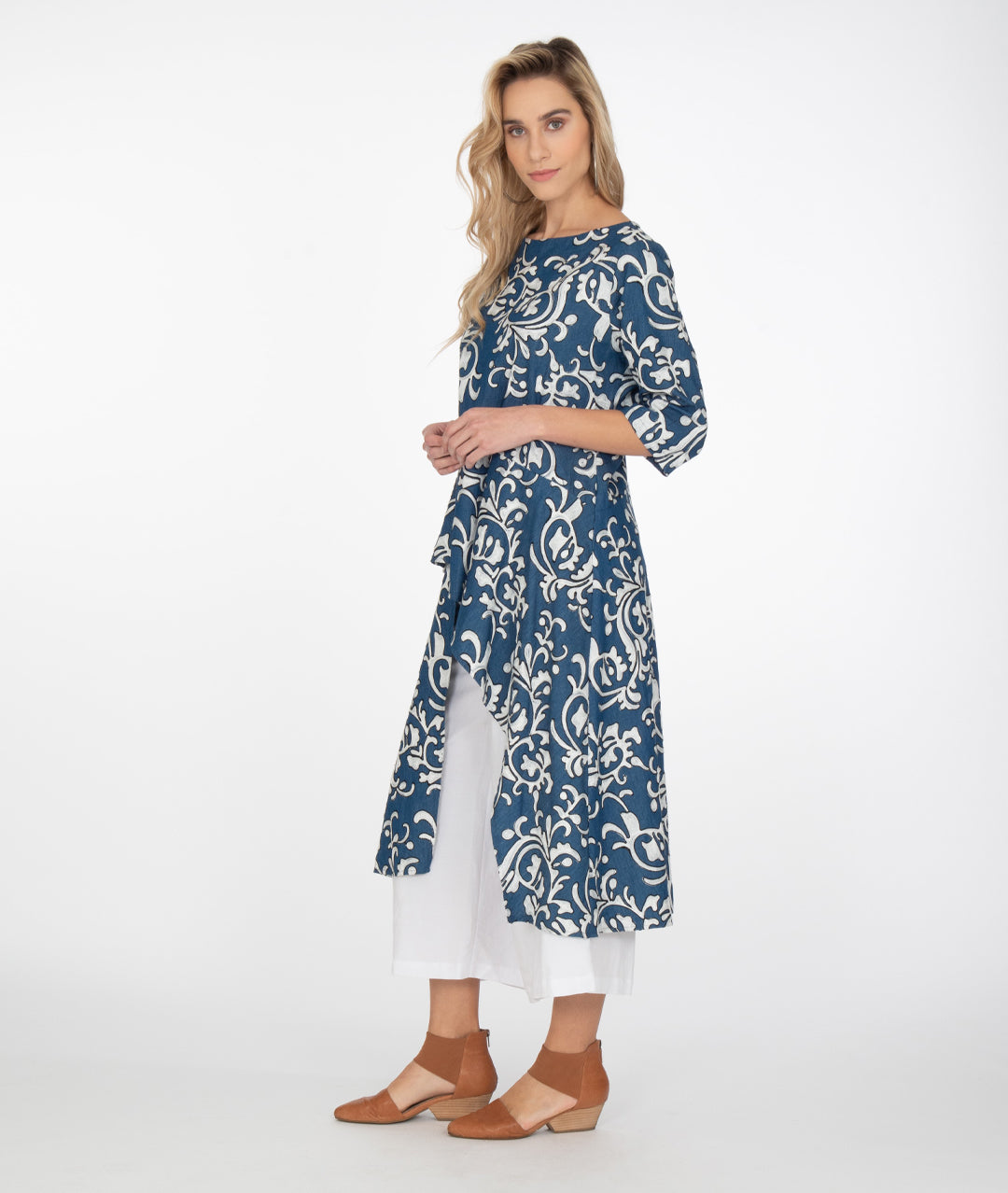 model in a wide leg white pant with a blue and white scroll print embroidered tunic with elbow length sleeves and an asymmetrical waist and skirt, with a short front and long sides and back. pockets set into side seams