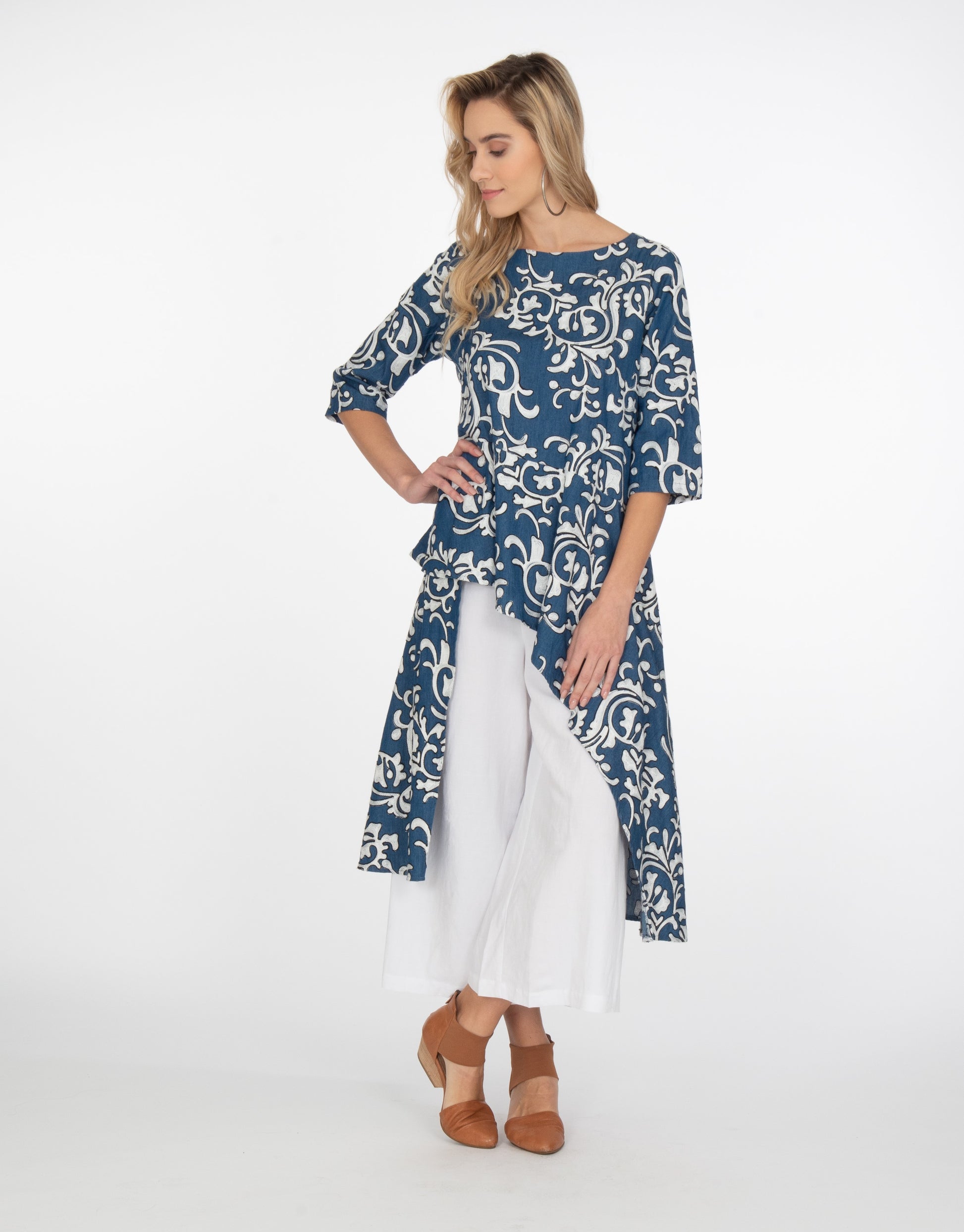 model in a wide leg white pant with a blue and white scroll print embroidered tunic with elbow length sleeves and an asymmetrical waist and skirt, with a short front and long sides and back. pockets set into side seams