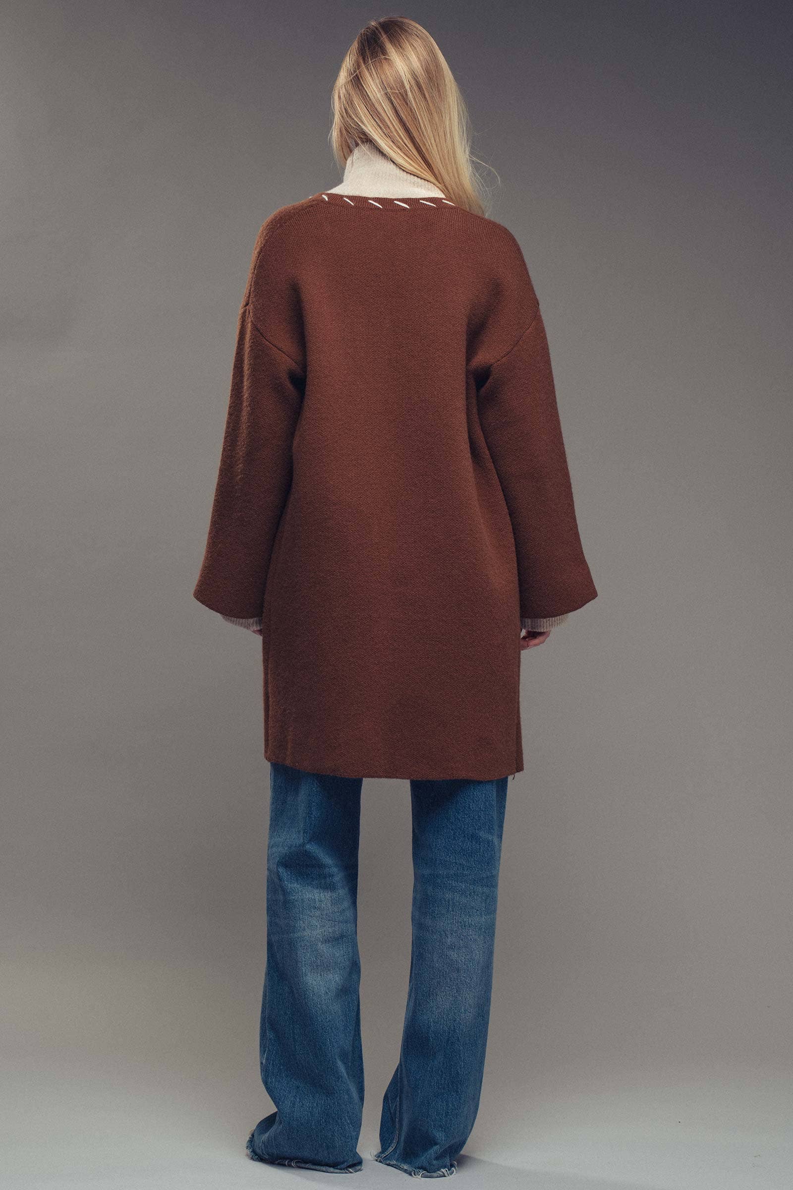 Woman wearing a brown coat with white stitching, beige turtleneck, and blue jeans on a gray background