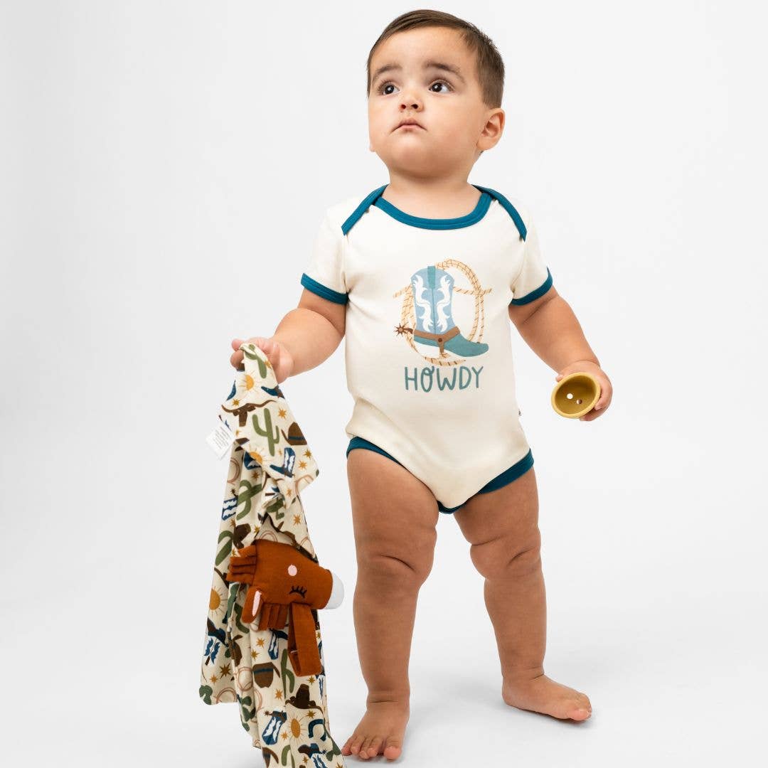 Baby wearing a 'Howdy' onesie with a patterned blanket and toy on a white background