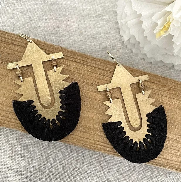 Pair of gold and black earrings on a wooden surface with white flowers in the background.