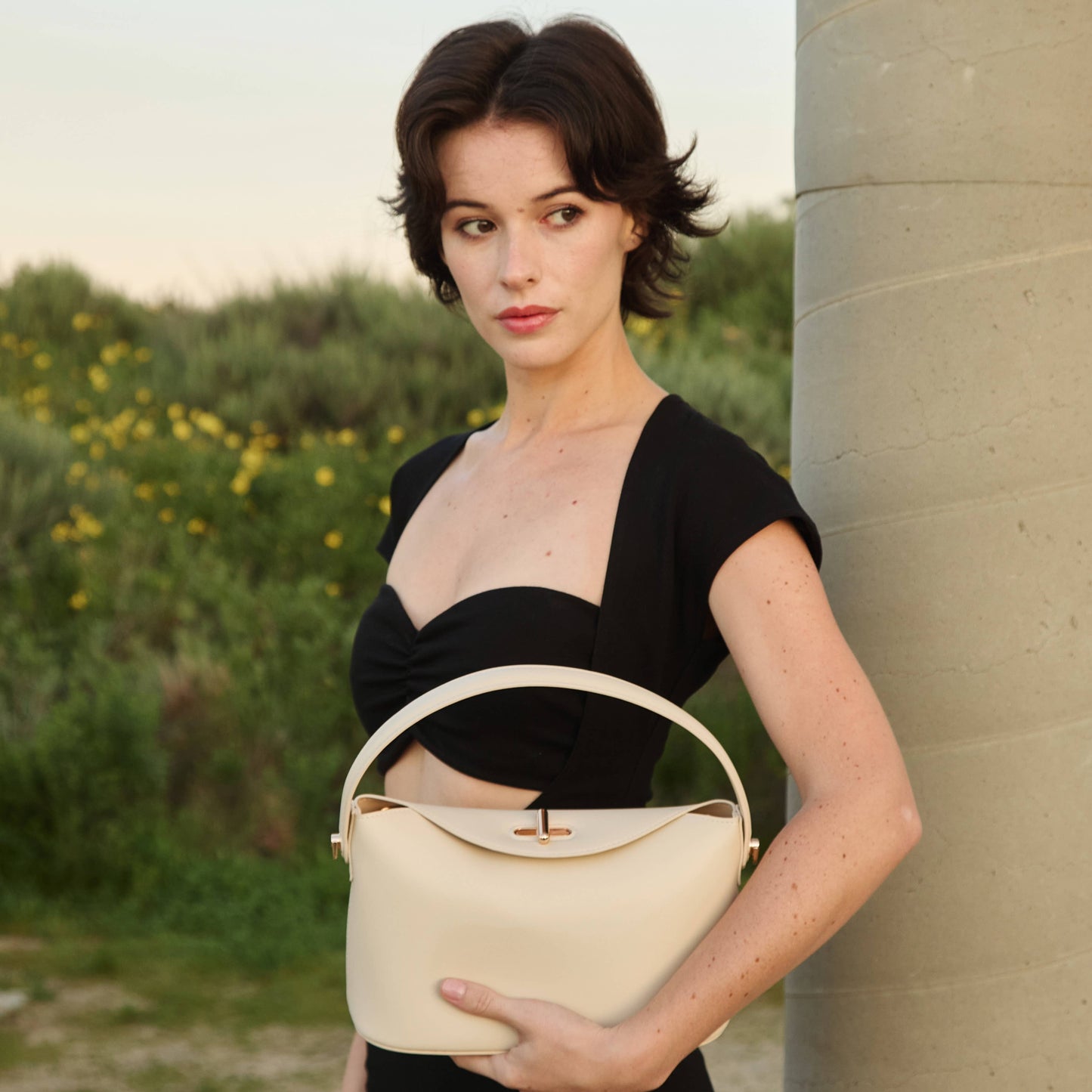 Woman holding a beige handbag in a natural setting with greenery.