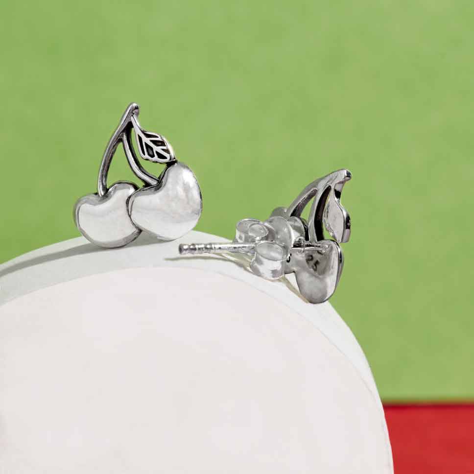 The image shows a pair of sterling silver cherry post earrings displayed on a white form, with a green background.
