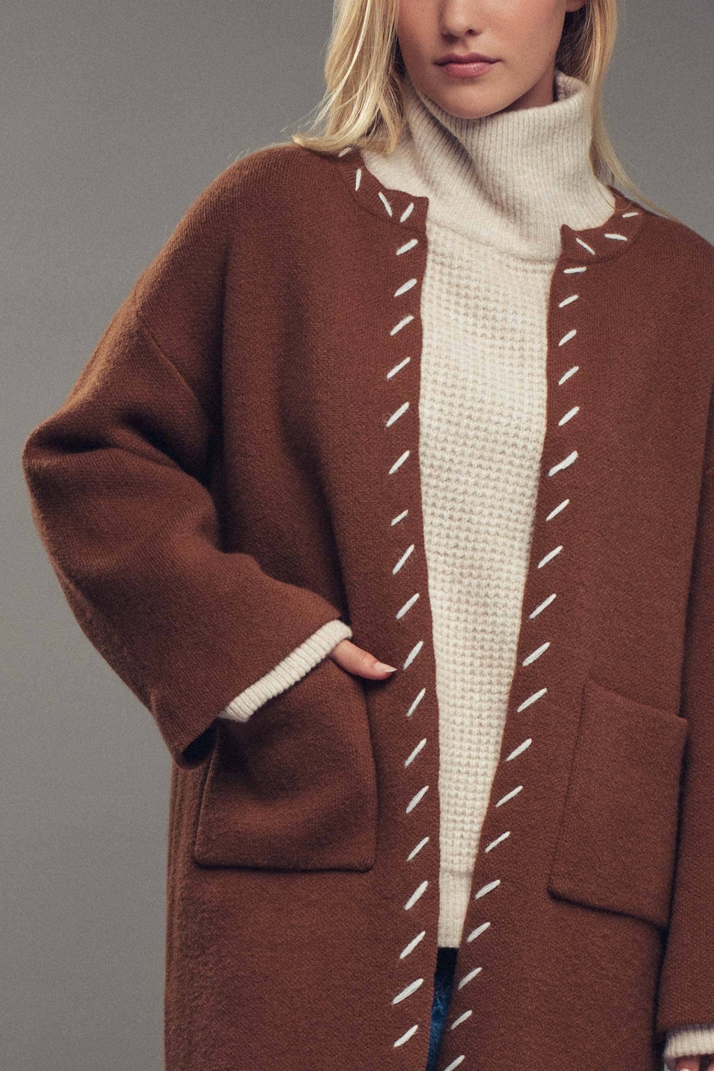 Woman wearing a brown coat with white stitching, beige turtleneck, and blue jeans on a gray background