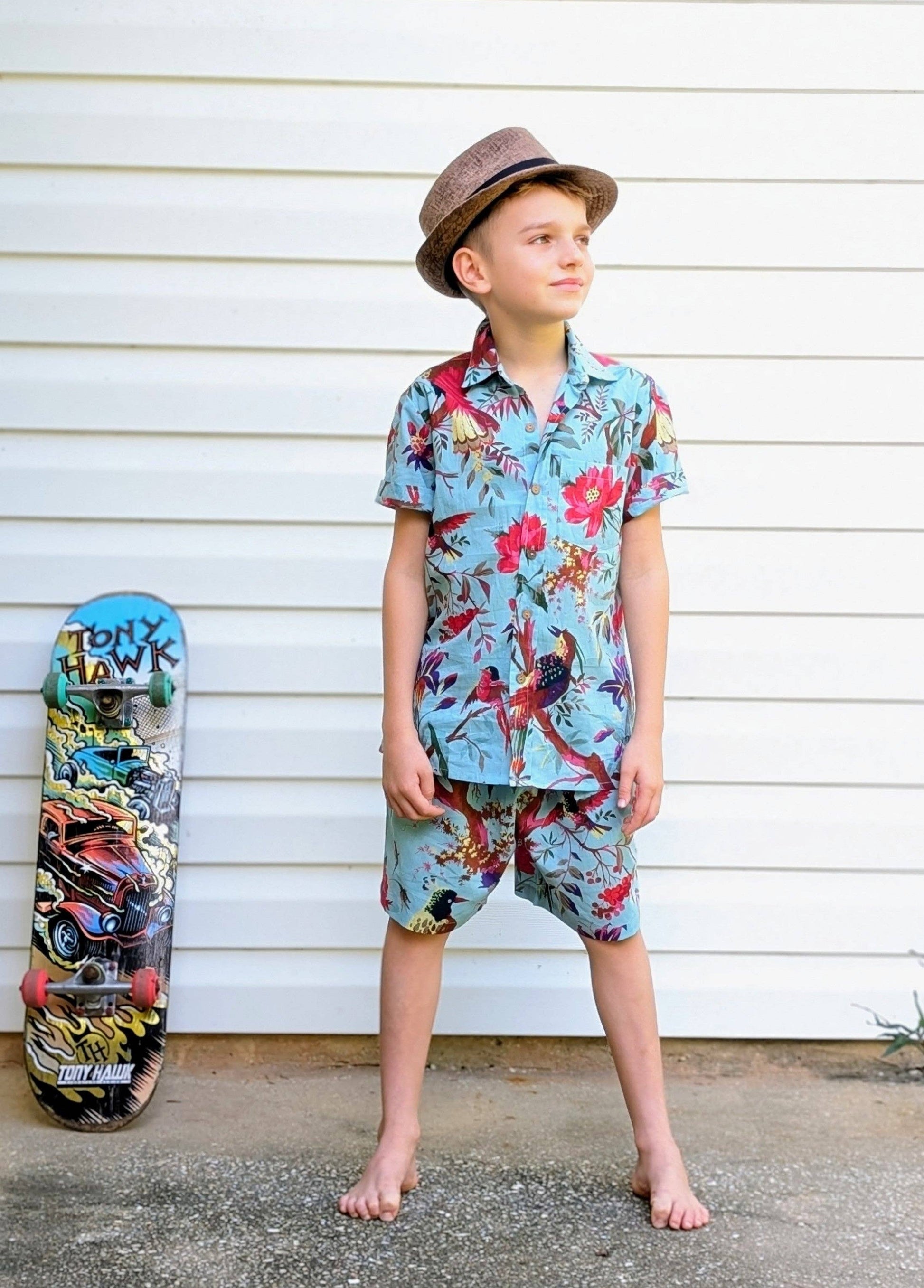 Child wearing a colorful floral shirt and shorts standing next to a skateboard against a white wall.