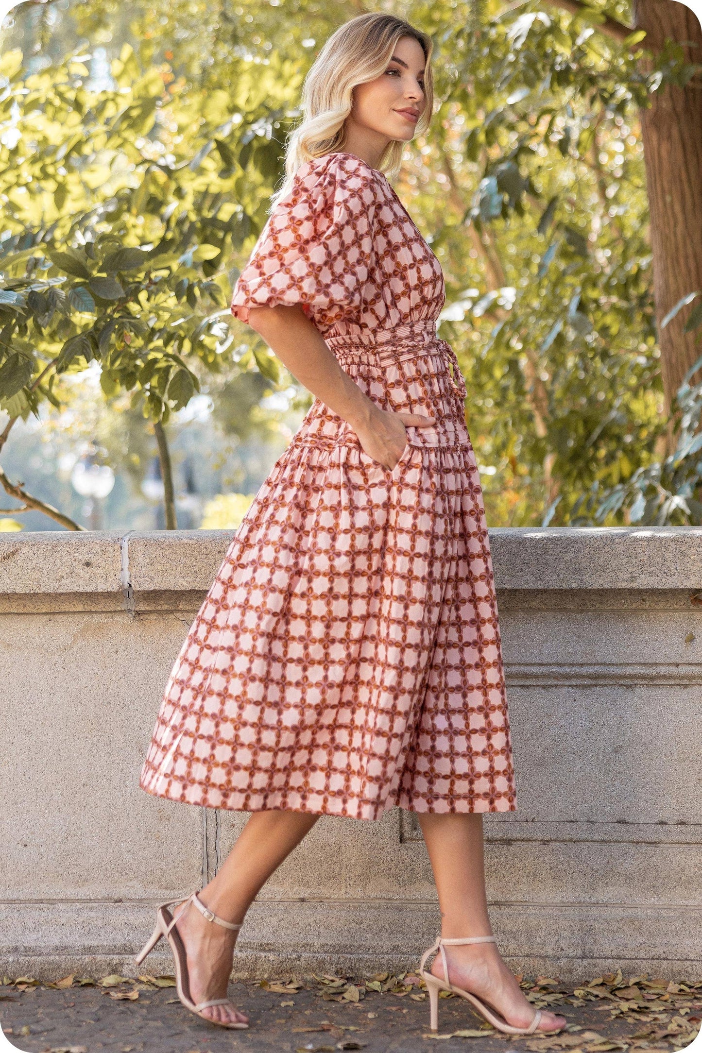 Woman wearing a red and white patterned dress standing outdoors with greenery in the background
