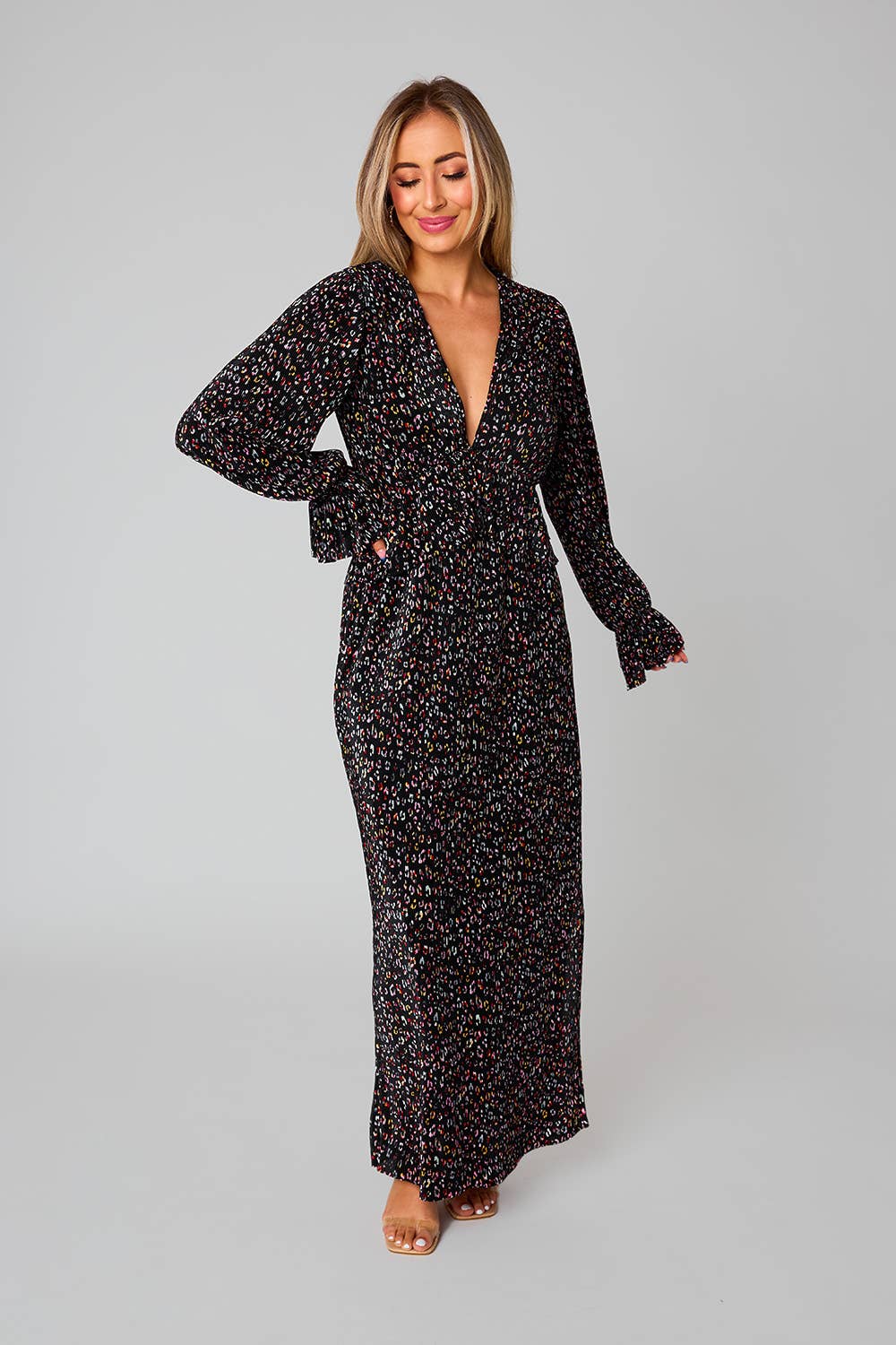 model wearing black long sleeve dress with elastic at the wrists for a flare wrist, plunging V neckline, a ruffle at the waistline, and a knee length slit on the side with colorful leopard print pattern throughout