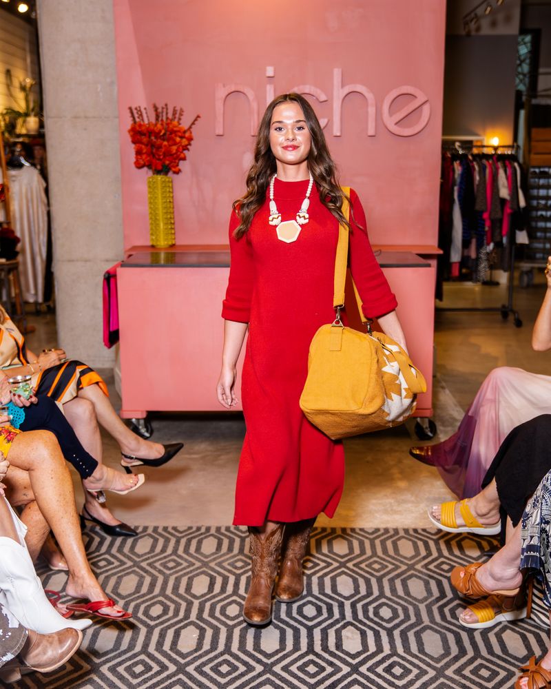 model wearing a red dress, chunky necklace and cowboy boots holding a yellow bag on a runway