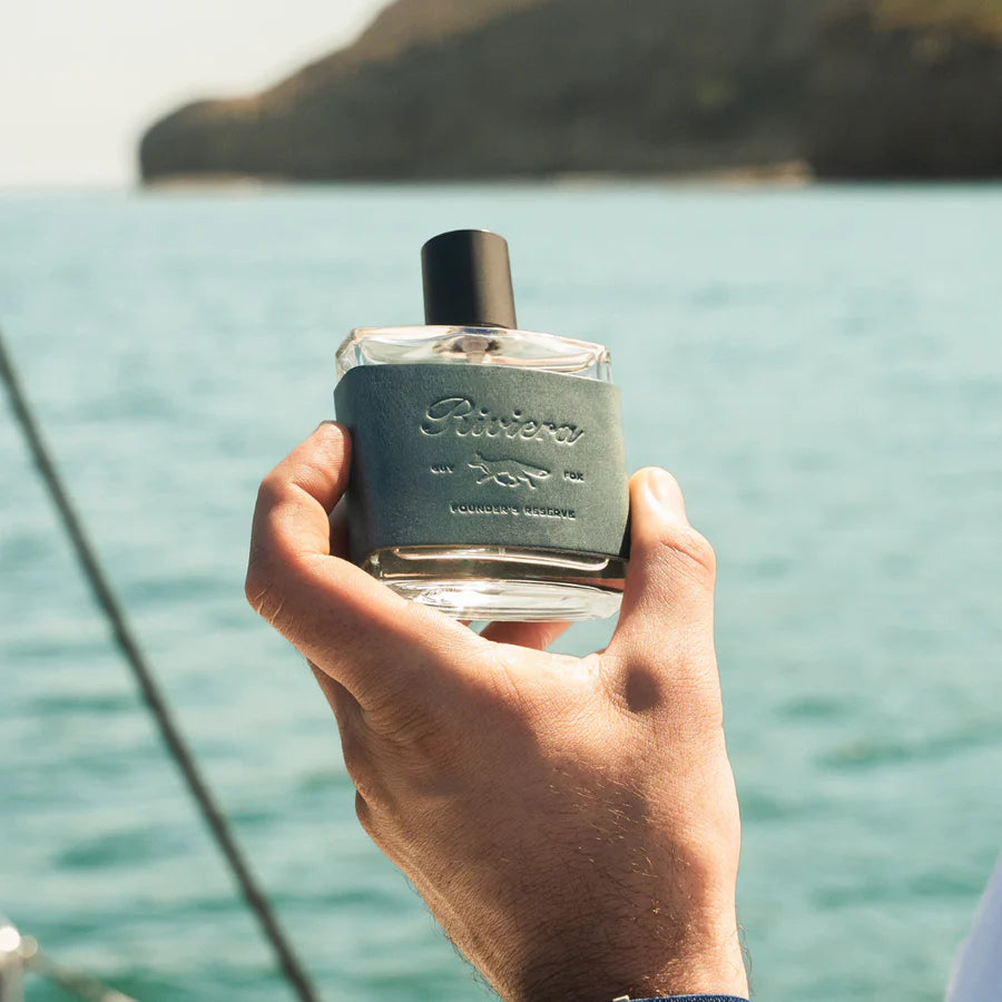 Hand holding a perfume bottle with a scenic background of water and sky.