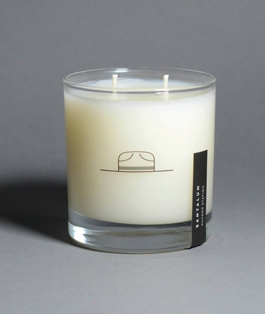 round glass candle with two wicks and small hat logo. small black label on the right side from bottom. on a grey background.