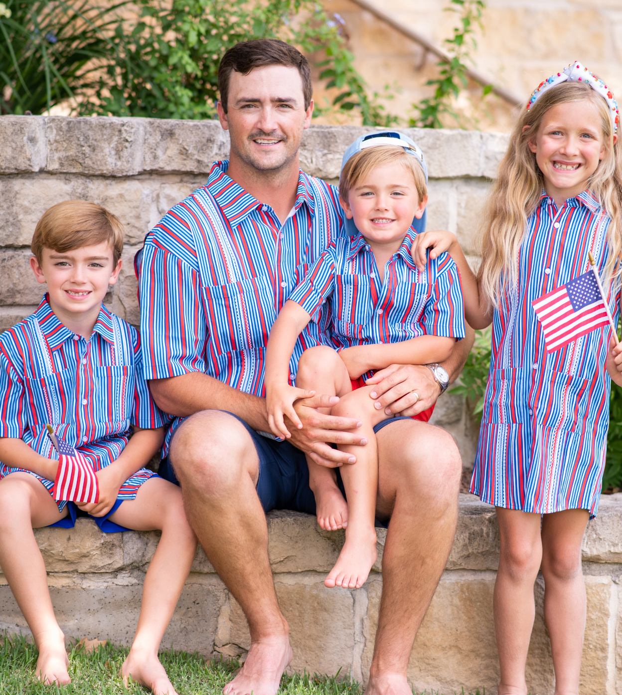 Man and three childeren sitting on a stone wall, dressed in the red, white and blue shirts.