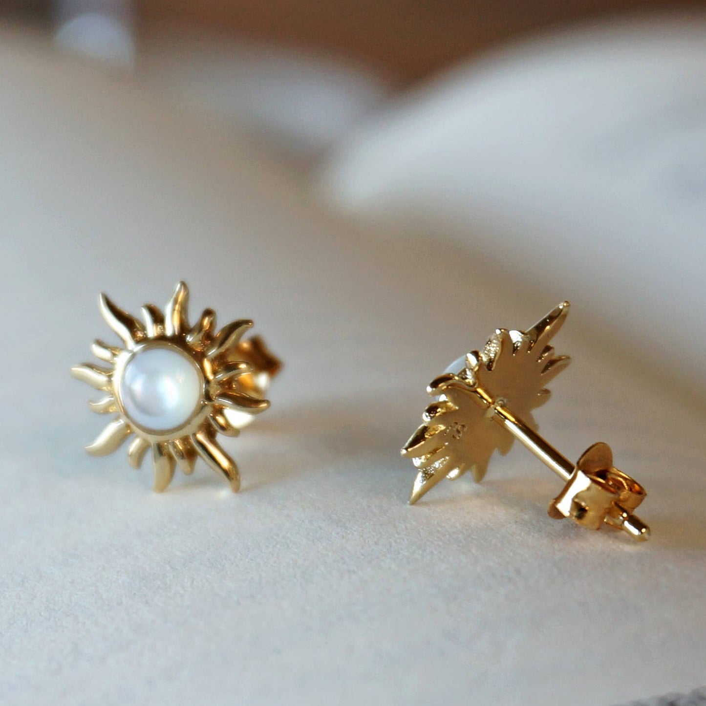 Gold sun-shaped earrings with a pearl center on a white surface.