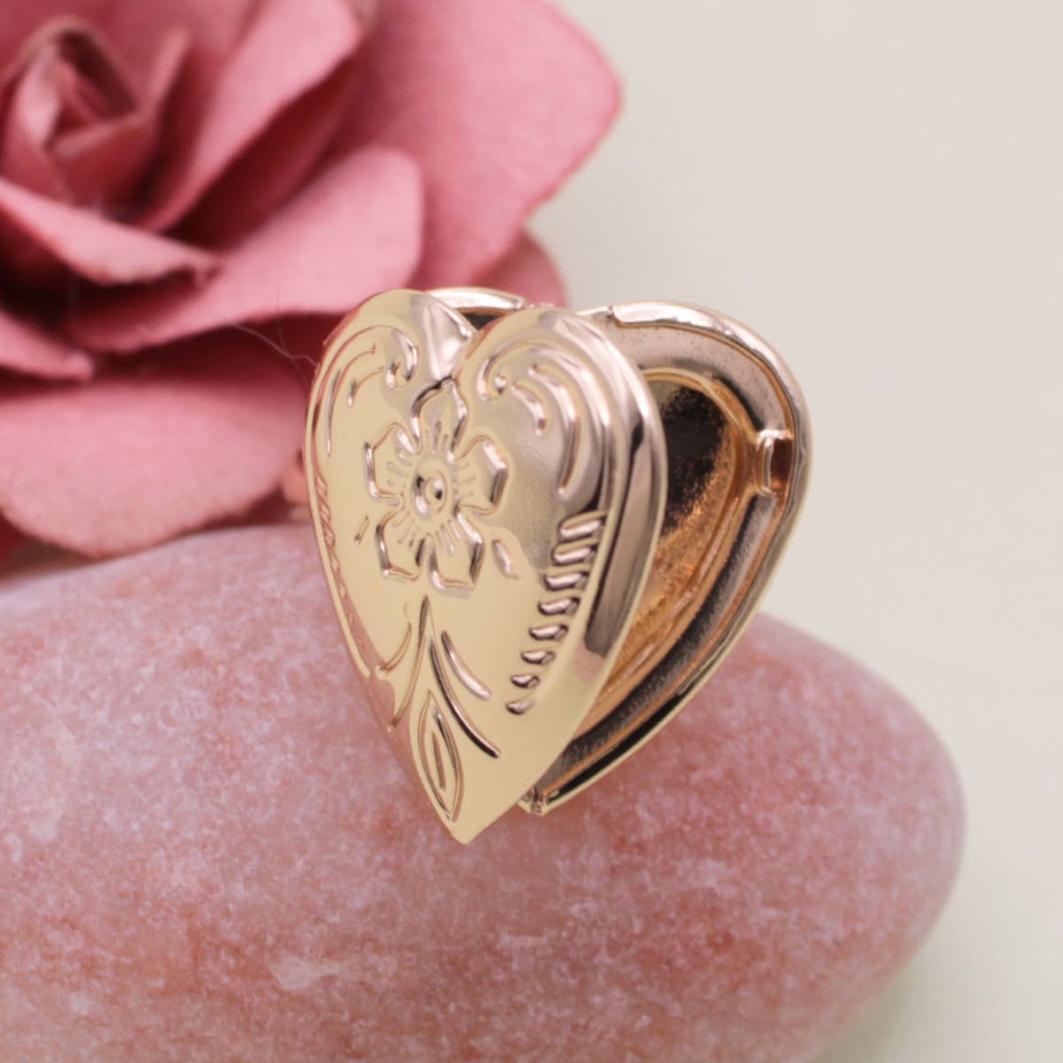 Gold heart-shaped locket ring on a pink stone with a blurred pink rose in the background
