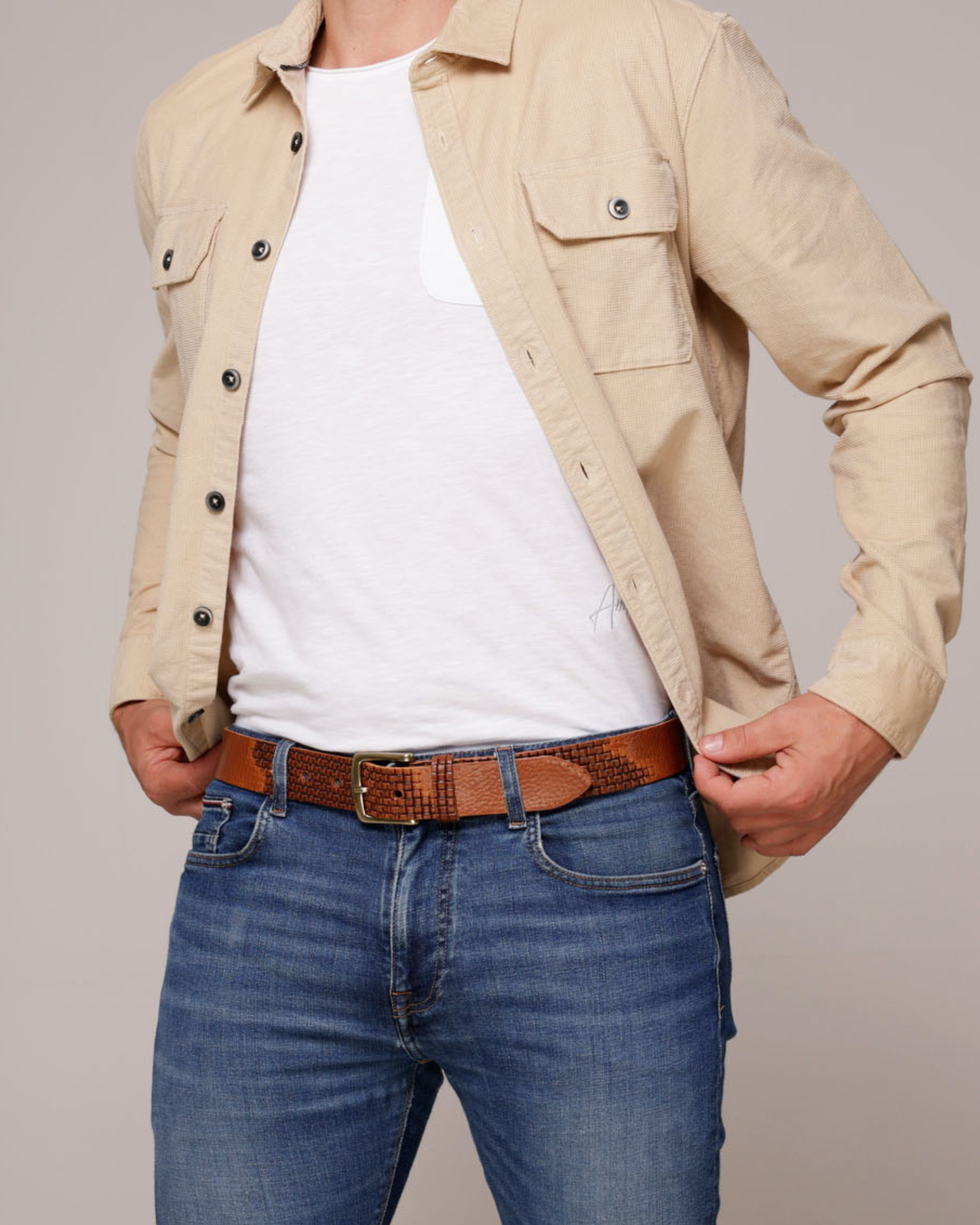 Person wearing a beige jacket, white shirt, and blue jeans with a brown belt on a plain background