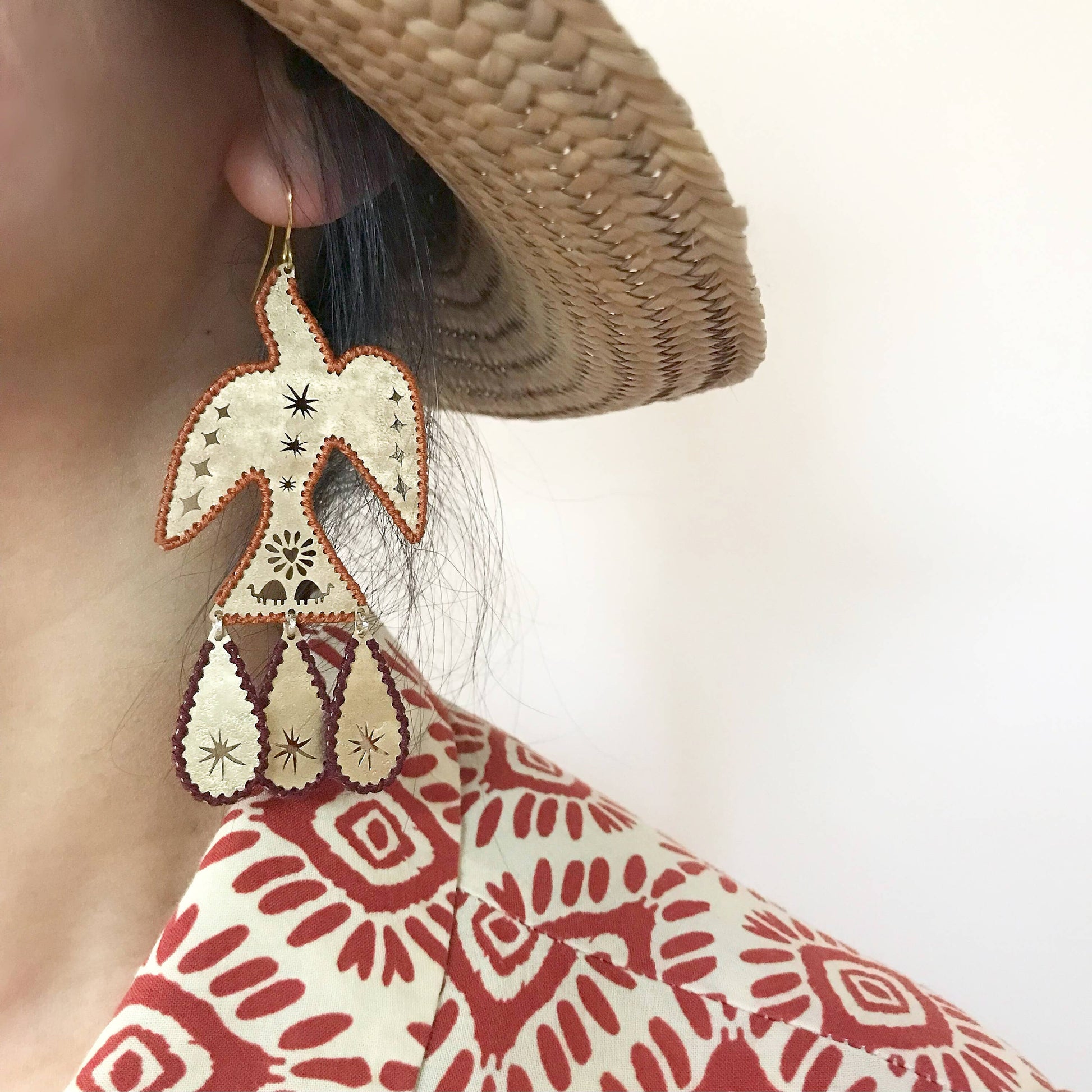Earring with geometric design worn by a person wearing a straw hat and patterned garment.