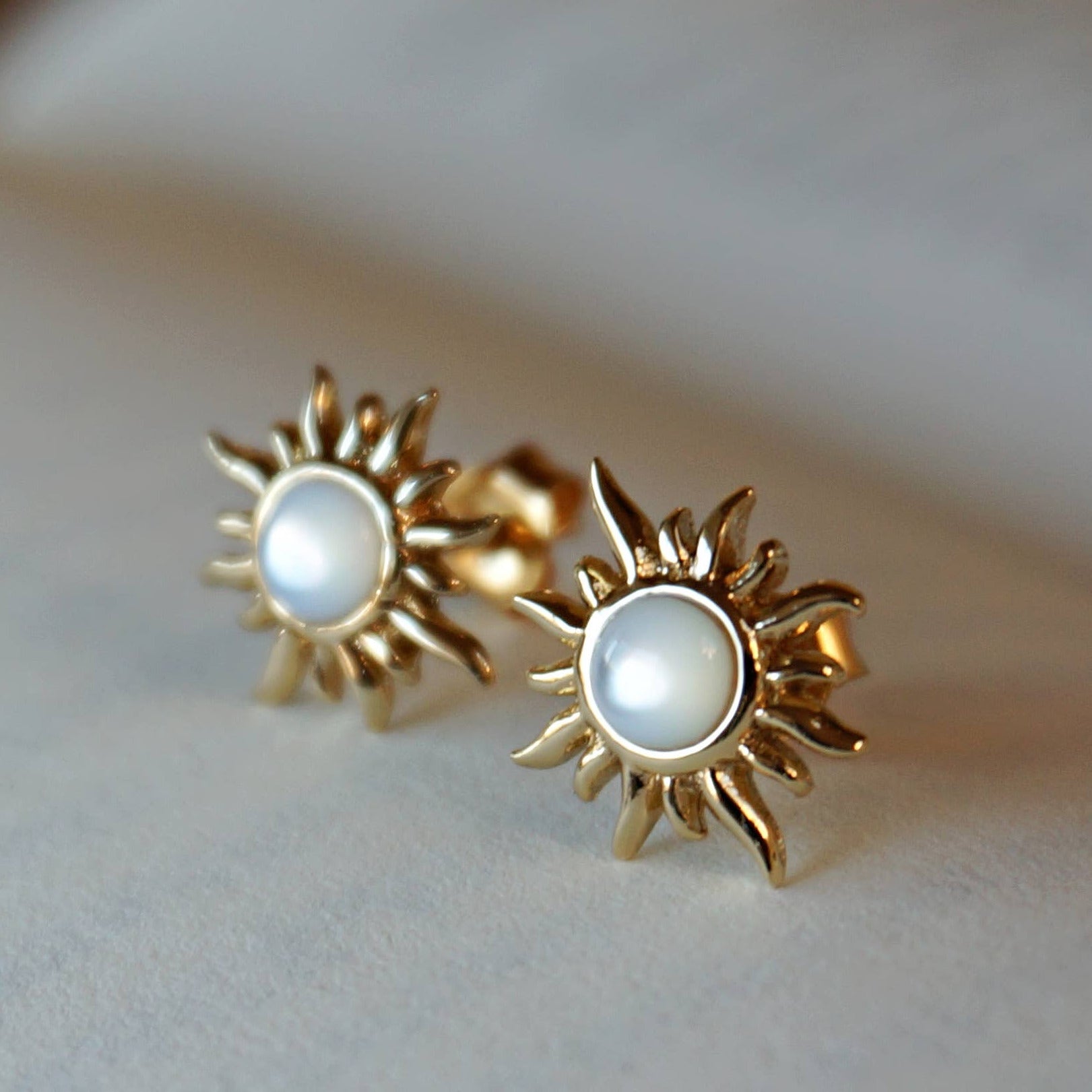 Gold sun-shaped earrings with pearl center on a soft background.