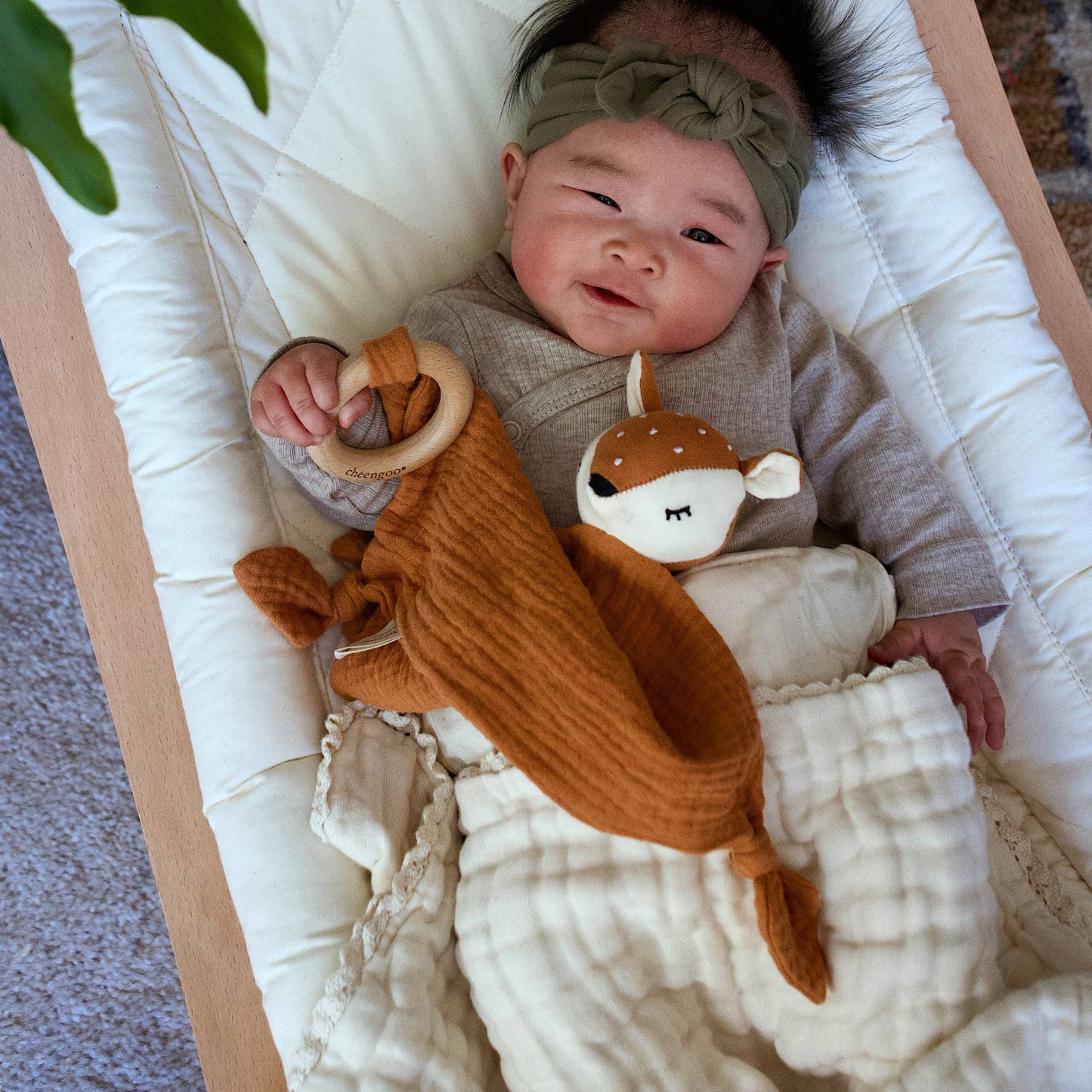 Baby lying on a white blanket with a brown plush toy, wearing a green headband.
