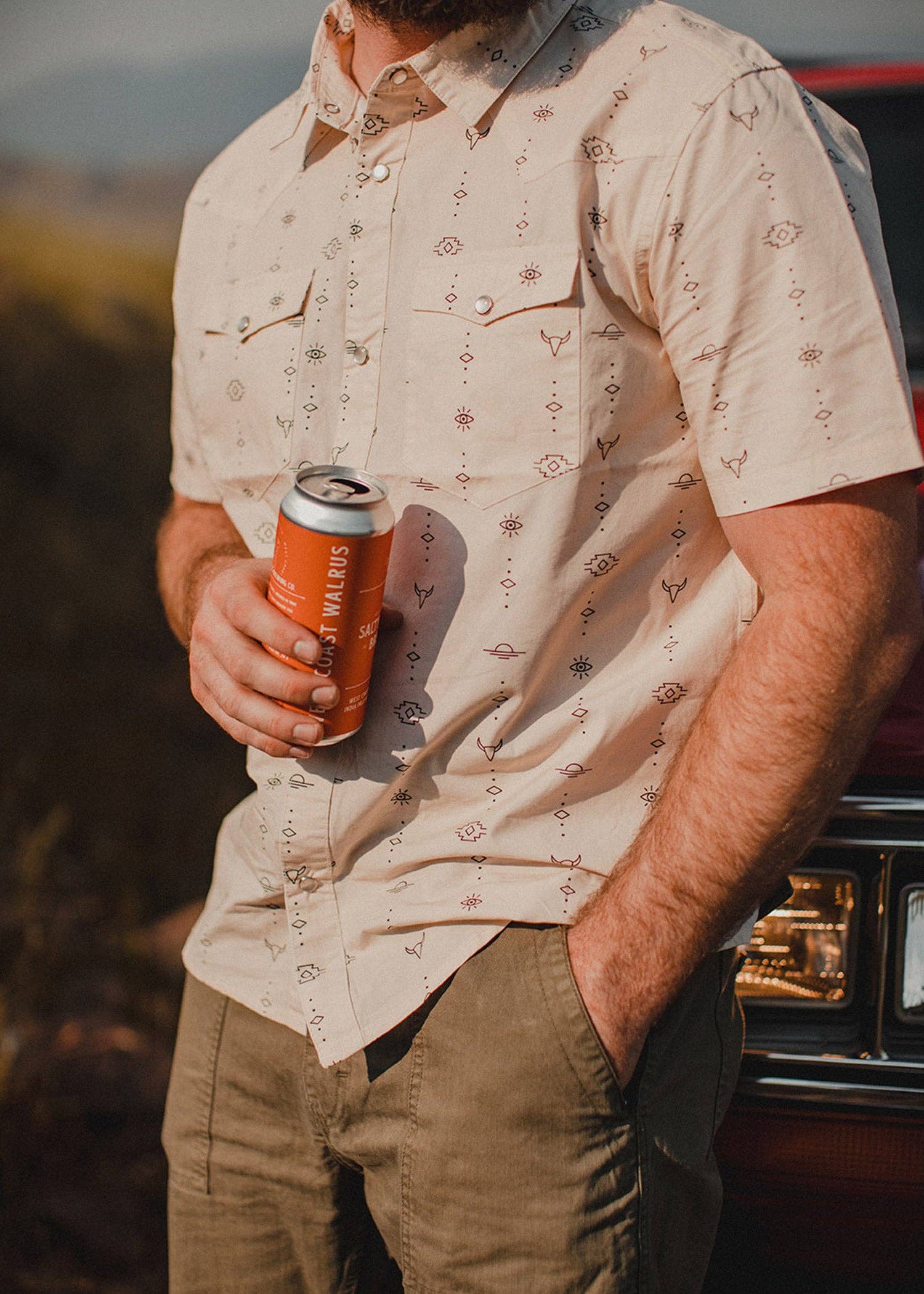 model wearing beige short sleeve shirt with line drawing print leaning on a car and holding a aluminum beverege can