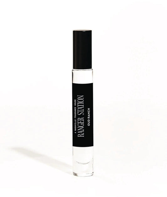 A small, cylindrical bottle of Oud Ranch Quickdraw Perfume with a black label and white cap, labeled with the brand's name and the scent 'Palo Santo / Oud Wood / Cedar / Pepper'.