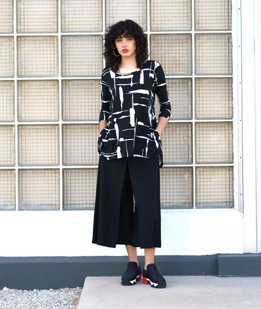 model wearing black and white print pullover top with a round neckline, 3/4 sleeves, and a wide hem with splits on either side, giving the illusion of a dipped hem with black pants and sneakers