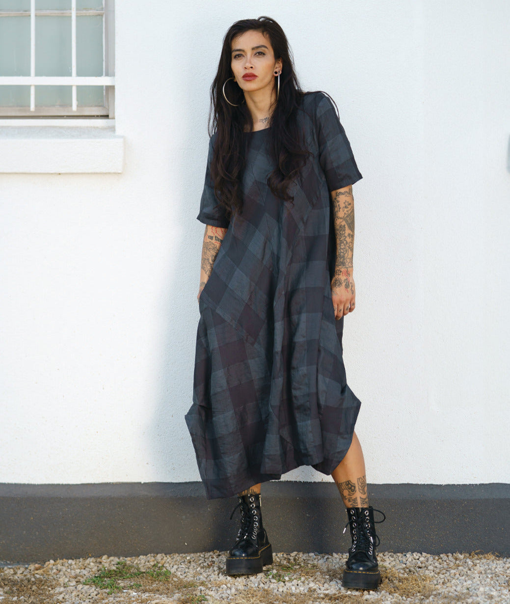 model in a midi length dress with short sleeves, a round neckline and tucks at the sides. dress has a large grid print on shades of deep gray with multi directional panels