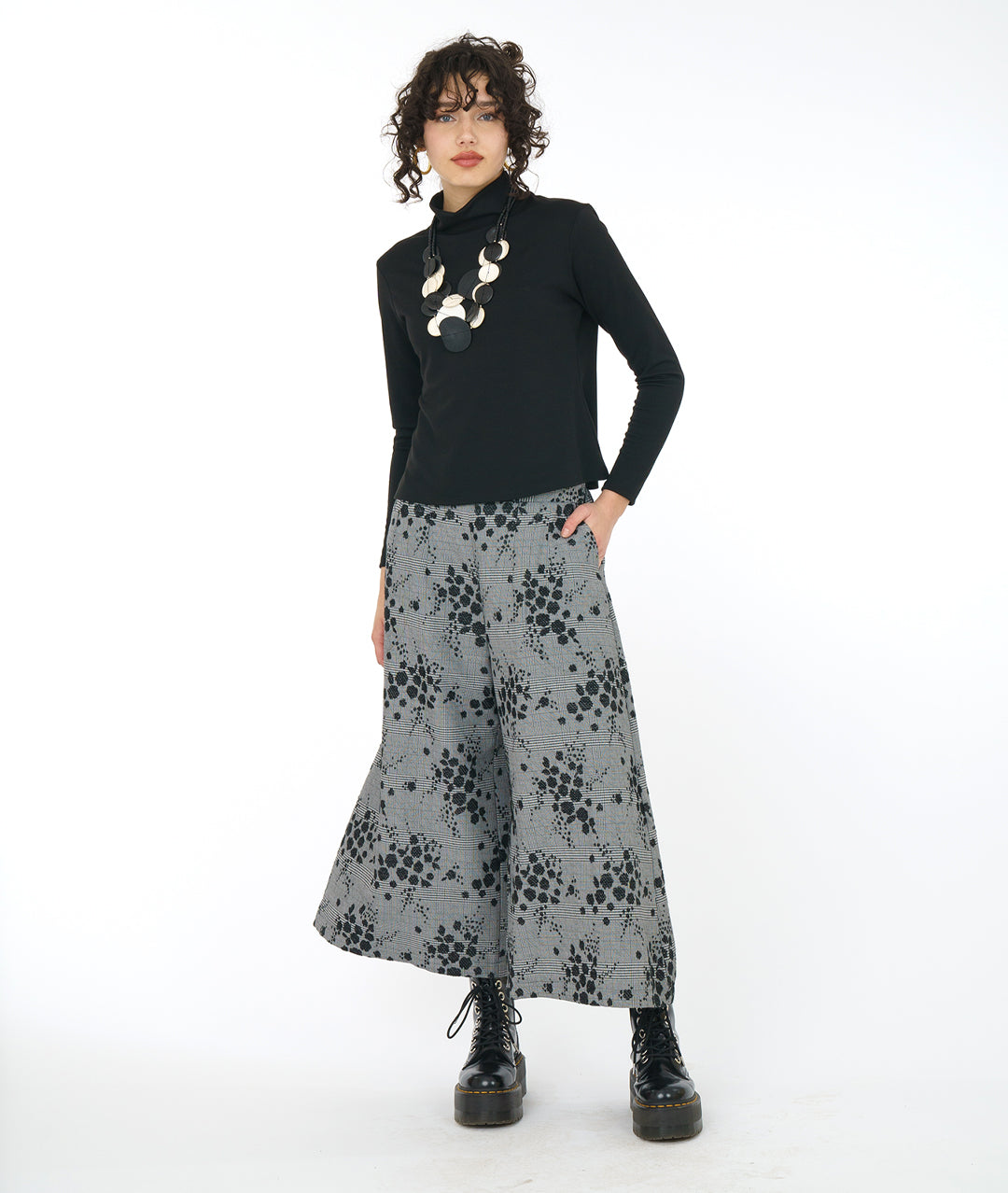 model in a black turtleneck and oversized chunky black and white necklace, worn with a cropped wide leg grey plaid pant with a black pollen print
